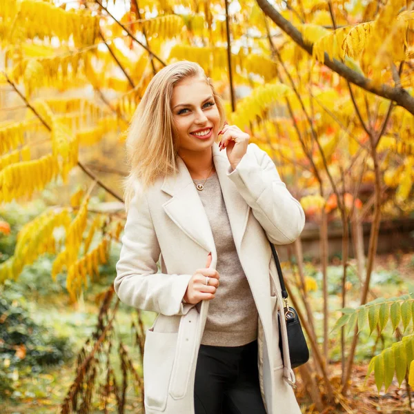 Красивая молодая счастливая женщина в стильном пальто с модной сумкой позирует возле желтых осенних листьев — стоковое фото