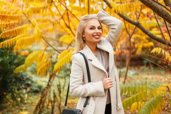 Stylowe piękna młoda kobieta z uśmiechem w modny płaszcz w pobliżu żółte liście jesienią — Zdjęcie stockowe