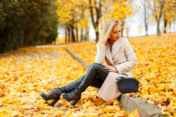 Mulher loira jovem e elegante bonita em um casaco moderno com sapatos elegantes e uma bolsa sentada no parque em um fundo de folhagem de outono — Fotografia de Stock