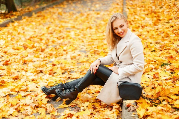 Schöne junge Frau in einem trendigen klassischen Mantel in eleganten Schuhen sitzt neben einem gelben Herbstlaub — Stockfoto