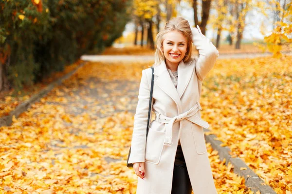 Feliz hermosa joven con una sonrisa en un abrigo clásico de moda con un bolso posando paseos en el día de otoño en un follaje amarillo — Foto de Stock