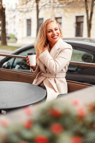 Happy beautiful young woman in trendy coat with coffee sitting on the street — Stock Photo, Image
