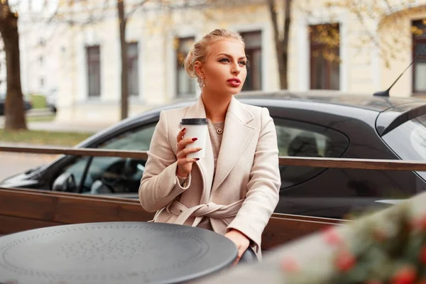 Стильная девушка с кофе в осеннем трикотажном пальто сидит за столиком на улице — стоковое фото