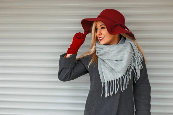 Mooie stijlvolle jongedame in een trendy jas met een grijze sjaal en elegante hoed poseren in de buurt van een houten muur — Stockfoto