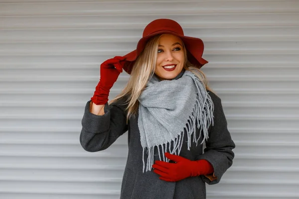 Elegante mooie jonge vrouw in een trendy hoed met een grijze sjaal in een stijlvolle vacht met handschoenen poseren in de buurt van een houten muur — Stockfoto