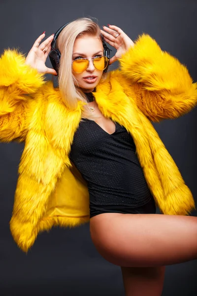 Élégante jeune femme à la mode avec écouteurs en lunettes de soleil jaunes et manteau en fourrure jaune posant en studio sur fond gris. Écoute de la musique — Photo