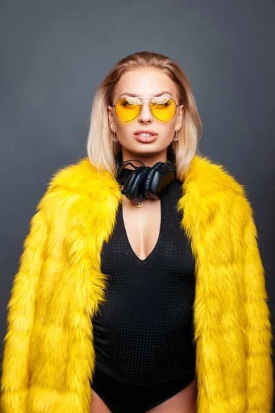 Hermosa mujer joven con auriculares gafas de sol con estilo de moda y abrigo de piel con estilo amarillo sobre un fondo gris —  Fotos de Stock