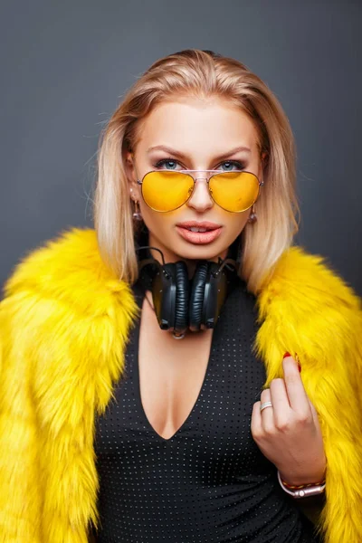 Jeune femme élégante à la mode avec des écouteurs avec des lunettes de soleil de mode jaune et manteau de fourrure jaune élégant sur un fond gris — Photo