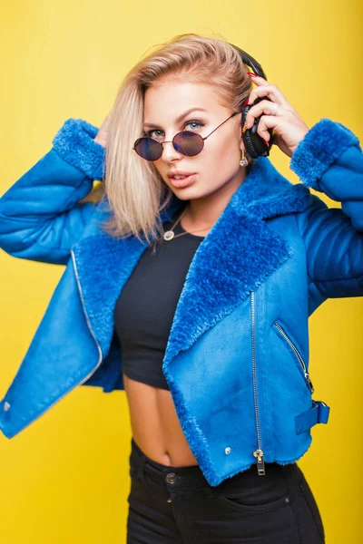 Chica joven con estilo en gafas de sol de moda con auriculares en una chaqueta de moda azul sobre un fondo. DJ mujer escuchando música —  Fotos de Stock