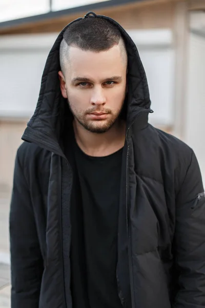Stylish beautiful young man with a hood in a black winter jacket on the street — Stock Photo, Image