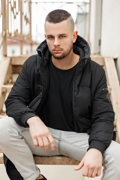 Hermosa joven modelo de un hombre con un corte de pelo corto en una chaqueta de moda de invierno se sienta en las escaleras — Foto de Stock