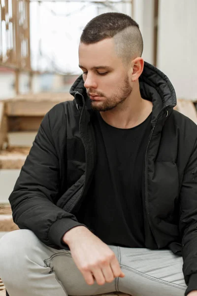 Knappe jonge man met een kort kapsel in een zwarte winter jas zit op de straat — Stockfoto