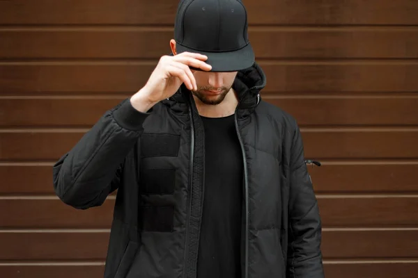 Νεαρός άνδρας σε ένα μαύρο μοντέρνο μπέιζ-μπώλ ΚΑΠ και ένα ζεστό μπουφάν χειμώνα ποζάρει στο δρόμο — Φωτογραφία Αρχείου