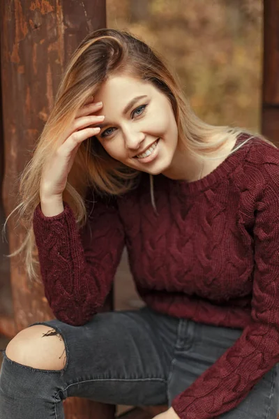 Mulher jovem feliz bonita Jovem feliz com um sorriso em uma camisola de malha elegante quente senta-se em um banco de madeira no dia de outono no parque com um sorriso em um tricô elegante quente. — Fotografia de Stock