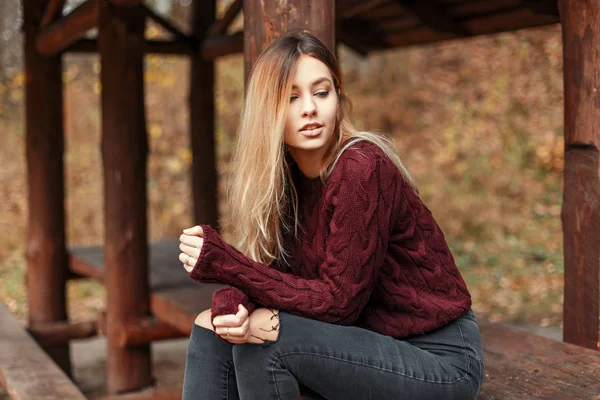 Стильна молода жінка в модному светрі і джинсах сидить на дерев'яній лавці в природі — стокове фото