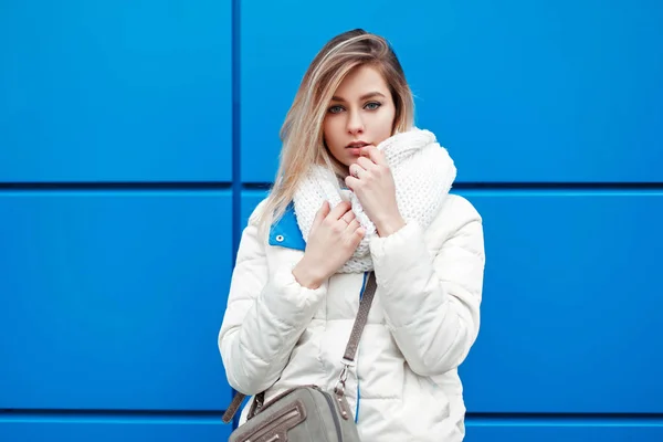 Красивая стильная молодая женщина с модным белым вязаным шарфом и зимней теплой курткой у голубой стены — стоковое фото