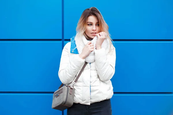 Mulher bonita na moda com um elegante lenço de malha branco e casaco de inverno branco com um saco de moda posando perto da parede azul — Fotografia de Stock