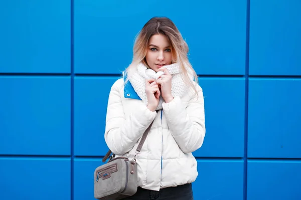 Jeune belle fille modèle en écharpe tricotée à la mode et veste d'hiver blanche avec sac à main posant près du mur bleu — Photo