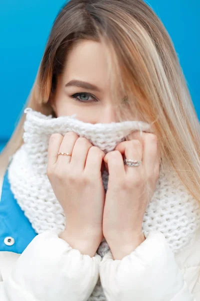 Retrato de una hermosa joven feliz con ojos azules con una bufanda de punto elegante y una chaqueta de invierno blanca sobre un fondo azul al aire libre — Foto de Stock