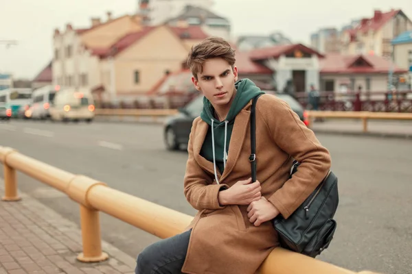 Knappe jonge stijlvolle man in een trendy jas met een hoodie met een stijlvolle tas zittend op de straat — Stockfoto