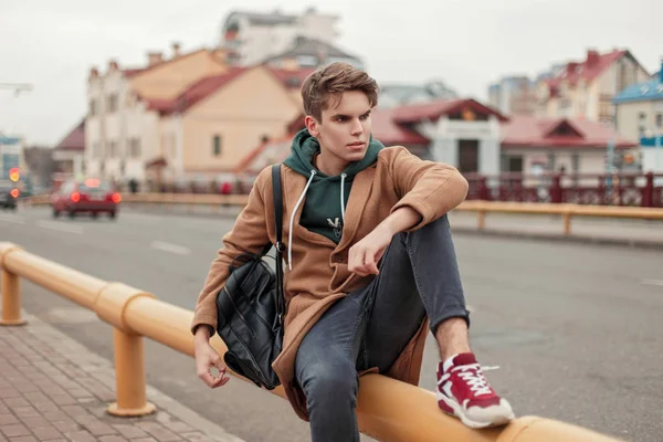 Jovem bonito com um corte de cabelo em um casaco vintage elegante com um saco preto de couro e tênis sentados perto da estrada na cidade — Fotografia de Stock