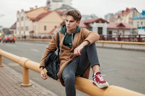 Chico modelo guapo de moda con un corte de pelo en un abrigo elegante vintage con una sudadera con capucha con una bolsa de cuero negro y zapatillas de deporte sentadas en una tubería en la ciudad — Foto de Stock