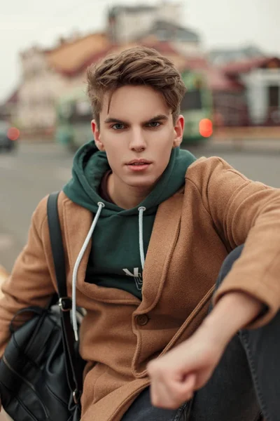 Joven de moda con un corte de pelo en un abrigo vintage con una sudadera con capucha y un bolso negro en la calle — Foto de Stock