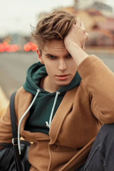 Joven hombre elegante con un corte de pelo en un abrigo de moda con una sudadera con capucha sentado en la calle — Foto de Stock