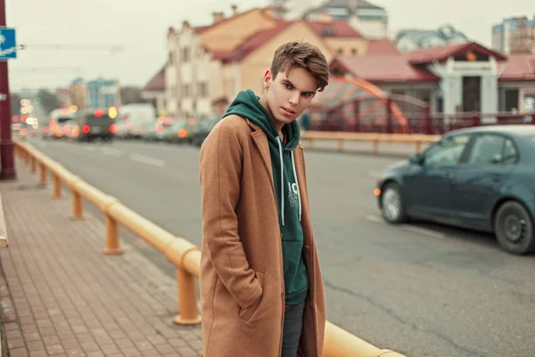 Guapo joven con abrigo y sudadera con capucha posando en la calle cerca de la carretera — Foto de Stock