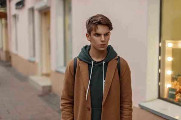 Snygga trendiga killen i en vintage kappa med en hoodie går på gatan nära butiker — Stockfoto
