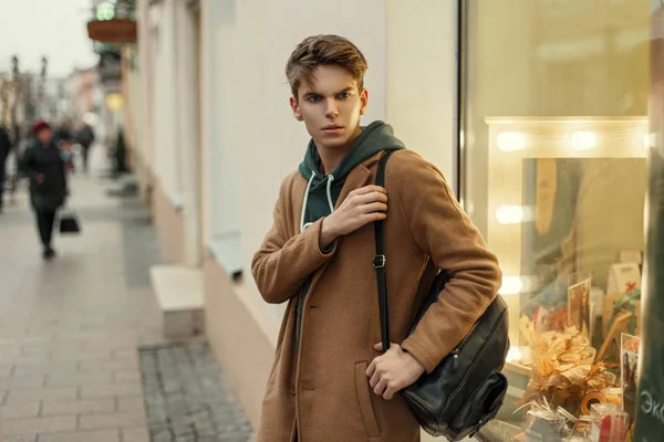 Joven guapo con un corte de pelo en un elegante abrigo de moda con una bolsa de cuero negro caminando al aire libre en la ciudad en un día de otoño — Foto de Stock