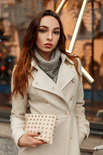 Mujer joven hermosa de moda en abrigo de moda con bolso elegante posando cerca de la ventana de la tienda en la calle — Foto de Stock