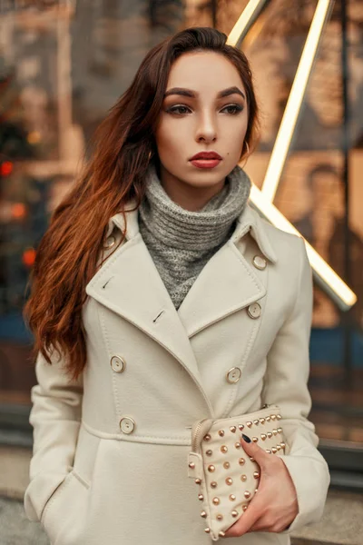 Jovem elegante em um casaco bege na moda com um saco elegante posando perto de uma janela de loja — Fotografia de Stock