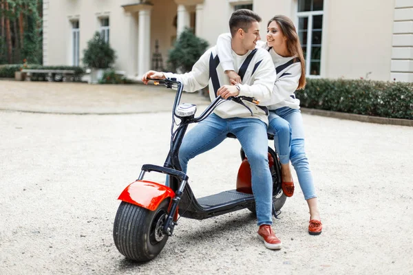 Homem europeu bonito com uma bela jovem mulher em roupas da moda está montando uma bicicleta elétrica perto do hotel. Casal apaixonado — Fotografia de Stock