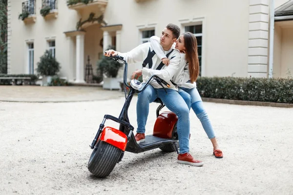 Homem bonito e uma mulher bonita na moda roupas elegantes com uma bicicleta elétrica perto do hotel — Fotografia de Stock