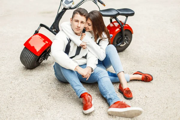 Casal bonito na moda em jeans roupas em sapatos vermelhos sentar e relaxar perto de uma bicicleta elétrica — Fotografia de Stock