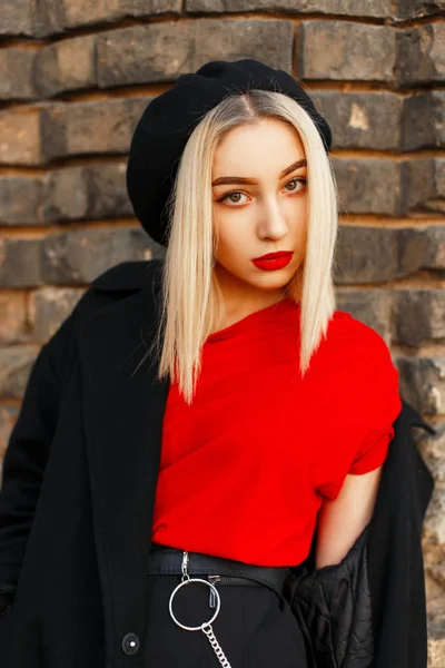 Retrato de una hermosa rubia con labios rojos en una boina negra y un abrigo negro cerca de una pared de ladrillo vintage —  Fotos de Stock