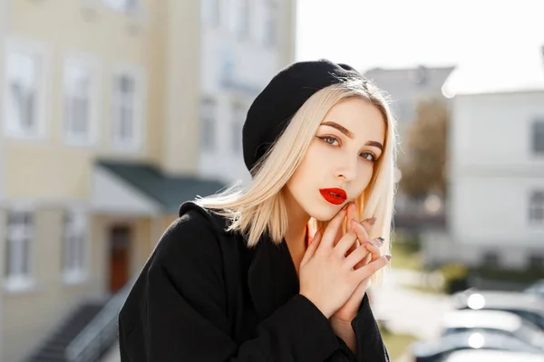 Ritratto alla moda di una bella donna bionda con un cappotto nero e un berretto nero in una giornata di sole — Foto Stock