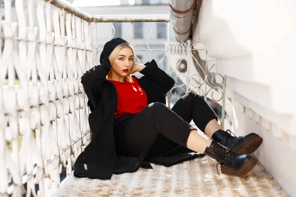 Modieuze jongedame in een stylish zwarte jas met een baret in laarzen zit in de buurt van de metalen reling — Stockfoto