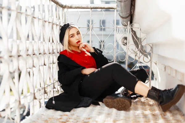 时髦的漂亮女人与红色的嘴唇在一个时髦的黑色大衣和贝雷帽与鞋坐在街上 — 图库照片