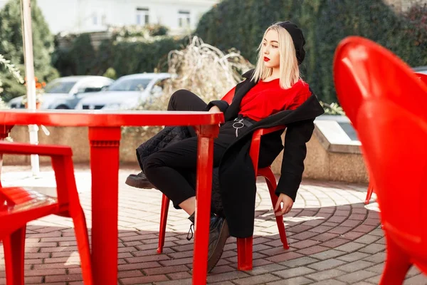 검은 코트의 유행가 옷에 젊은 아름 다운 여자는 카페에 빨간의 자에 앉아 — 스톡 사진