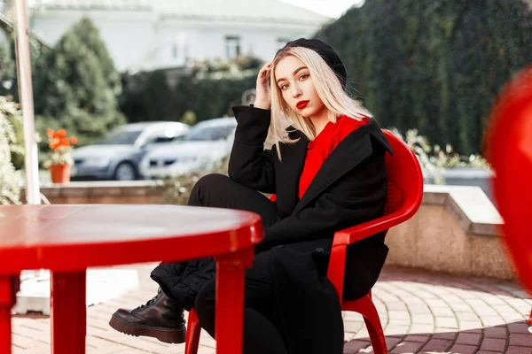 Élégante belle femme en manteau noir avec un béret assis sur une chaise rouge — Photo