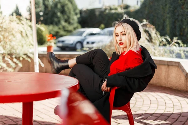 Belle femme à la mode dans un manteau noir avec un béret repose sur une chaise rouge — Photo