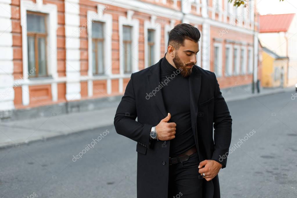 handsome stylish strong man in a black trendy coat walks on the street