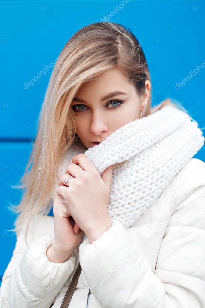 Young american model girl with fashionable white knitted scarf in a winter warm jacket near a blue wall on a winter day
