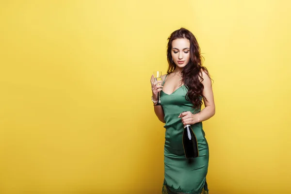 Élégante belle jeune fille dans une robe de soirée tendance avec une bouteille et un verre près du mur jaune — Photo
