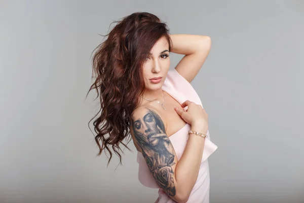 Retrato de moda de uma bela jovem com um corte de cabelo com uma tatuagem no braço em um vestido rosa elegante em um fundo cinza — Fotografia de Stock