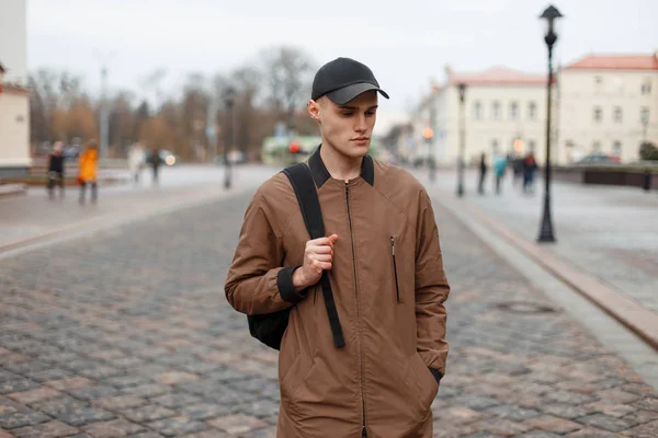 Joven hombre con estilo en un abrigo de moda de moda con una gorra y una espalda — Foto de Stock