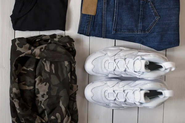 Vita sneakers, militära trasa, en svart T-shirt och blå jeans på ett trägolv. Fashionabla snygga mannens 2018 samlingar på en vit trä bakgrund. Ovanifrån — Stockfoto