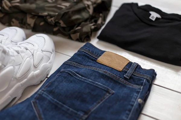 Coleção de verão masculino de roupas de moda em um fundo de madeira branca. Tênis, camiseta, tecido militar e jeans — Fotografia de Stock